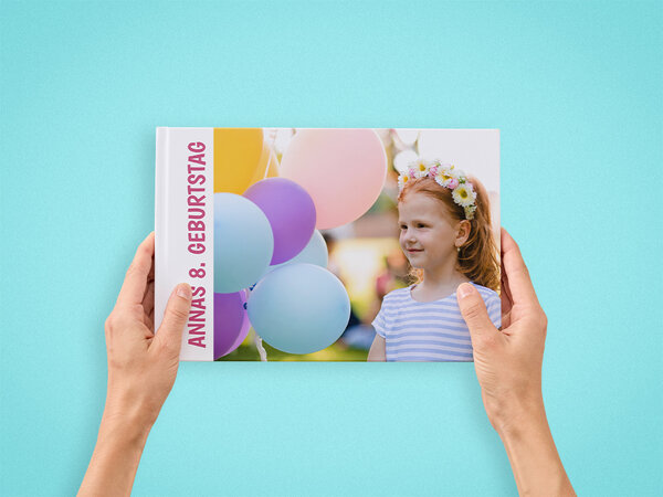Zwei Hände halten ein Fotobuch im A4-Querformat. Auf dem Cover ist ein Mädchen mit Luftballons im Hintergrund zu sehen. 