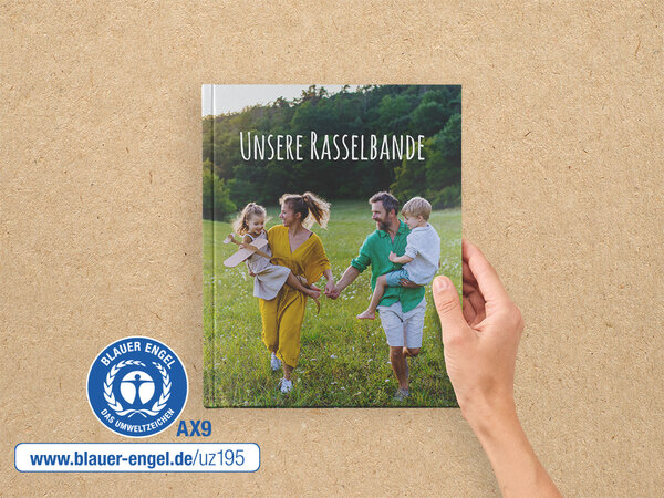 Eine Hand hält ein A4 Hoch Fotobuch. Auf dem Cover ist eine Familie auf einer Wiese abgebildet. Daneben ist der Blaue Engel.