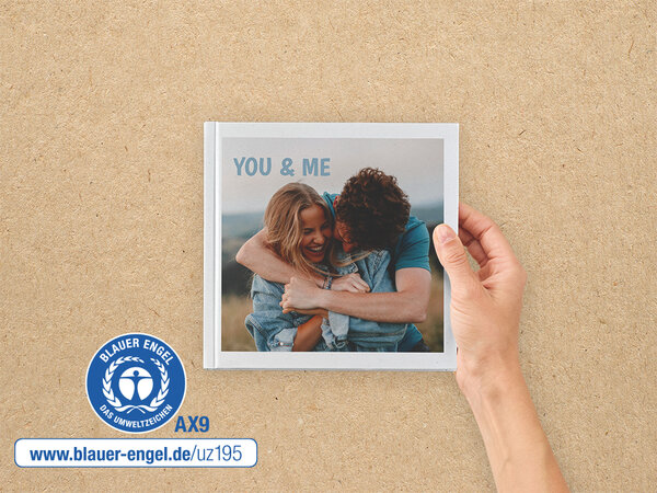 Ein Fotobuch im Format 20x20 wird von einer Hand gehalten. Auf dem Cover ist ein Paar zu sehen. Daneben ist der Blaue Engel.
